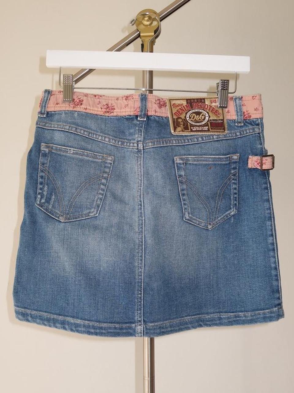 A blue denim d&g mini skirt shown from the back, hanging on a white hanger with a gold stand against a light beige wall. The back features two large patch pockets, a pink floral waistband lining, and a brown leather brand patch.
