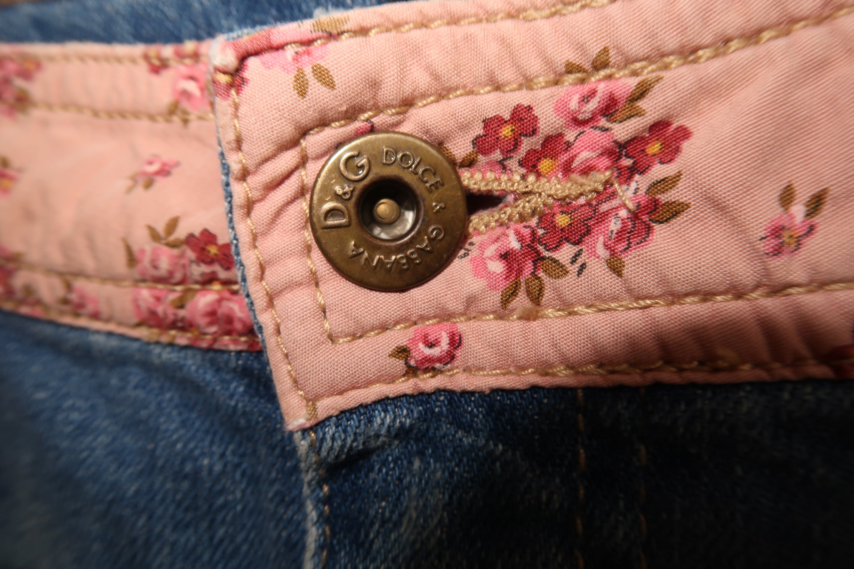 Close-up of a D&G Dolce & Gabbana pair of jeans showing the floral pink quilted waistband, a brass button with 'D&G Dolce & Gabbana' engraved, and blue denim fabric.