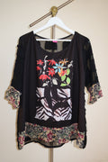 Save the queen y2k archive black sheer tunic top with a colorful floral and zebra print on the front, featuring patterned ruffled trim at the sleeves and hem, hanging on a white hanger against a beige wall.