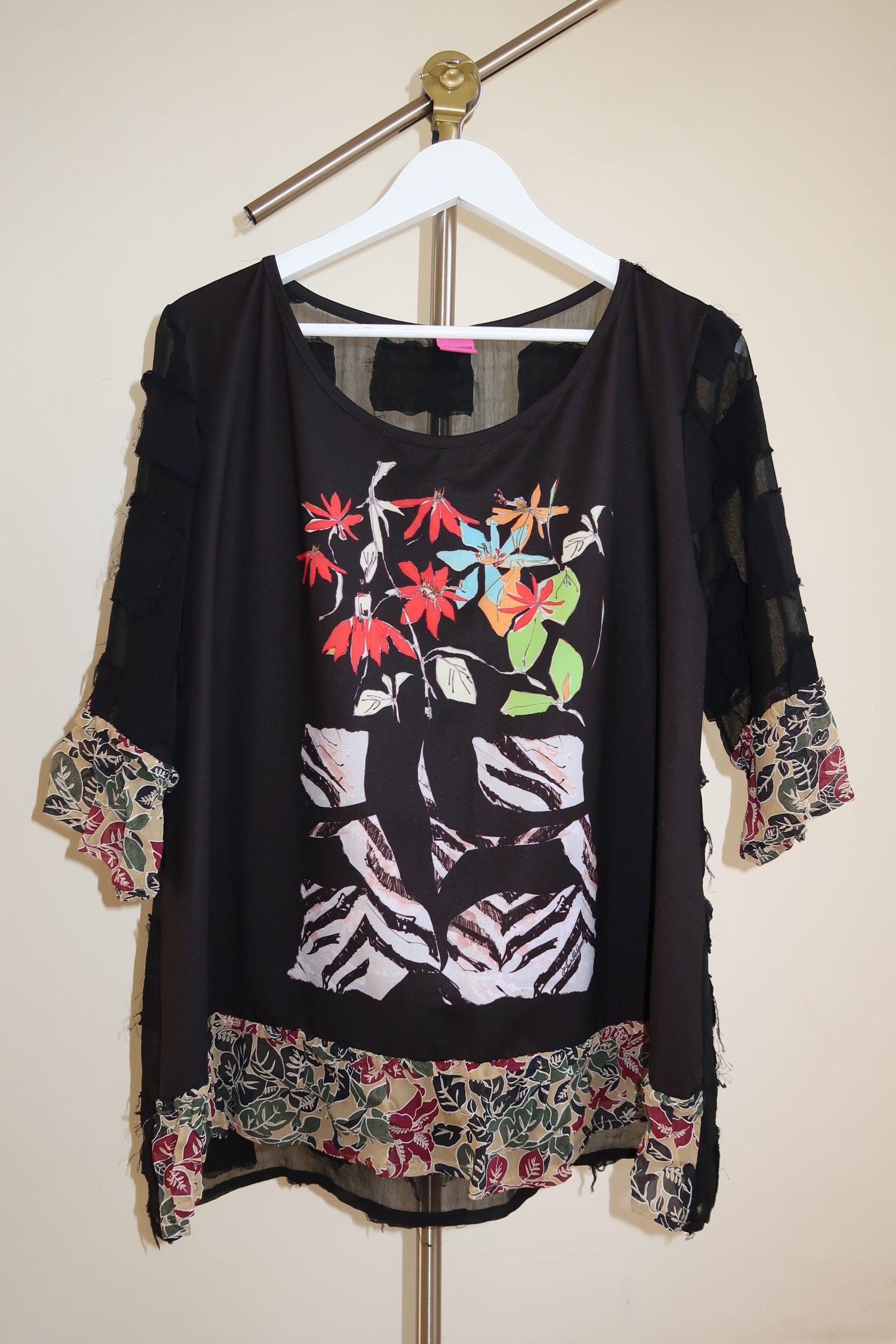 Save the queen y2k archive black sheer tunic top with a colorful floral and zebra print on the front, featuring patterned ruffled trim at the sleeves and hem, hanging on a white hanger against a beige wall.