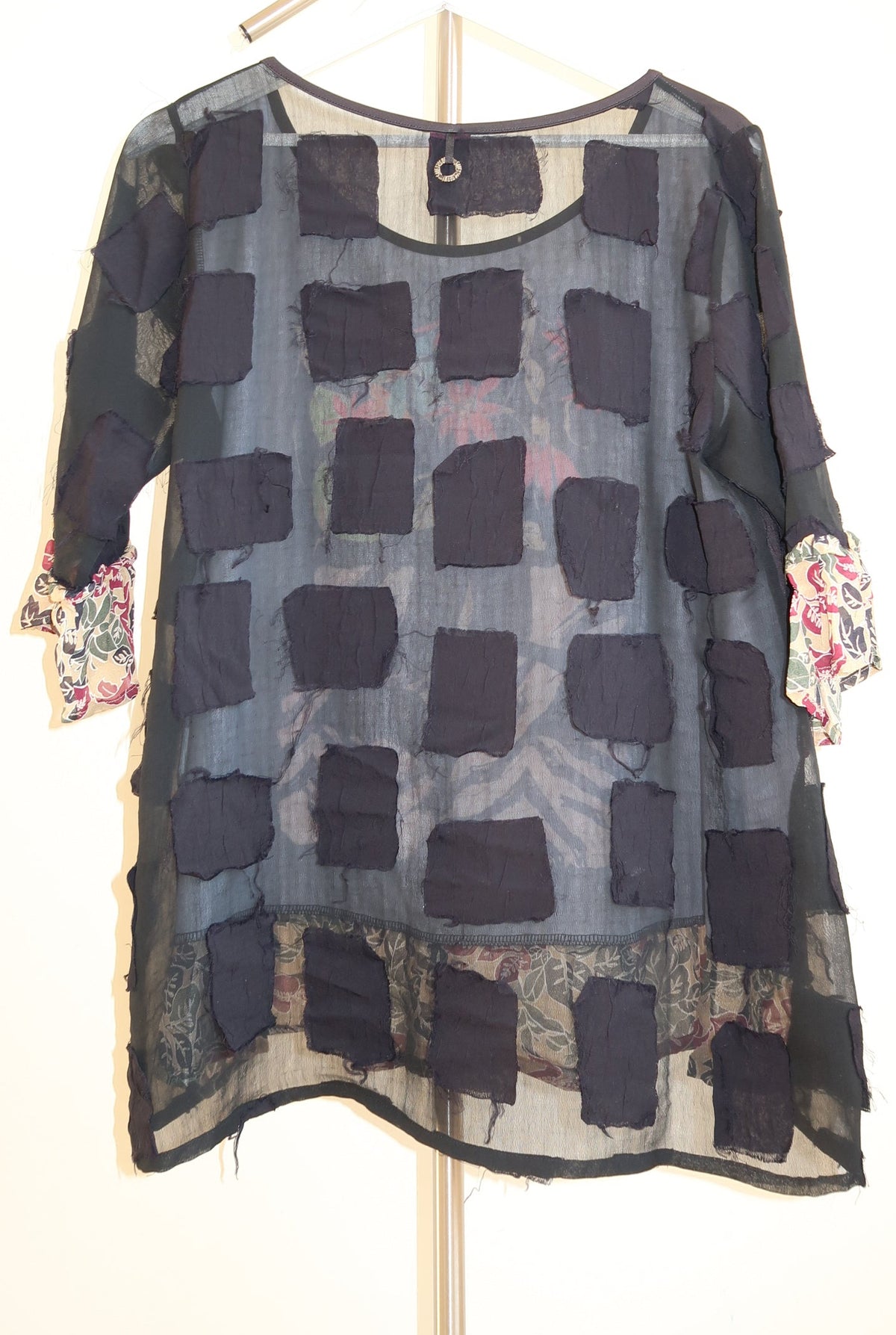 A sheer black patchwork tunic top with dark charcoal square patches, displayed on a hanger against a plain off-white background.